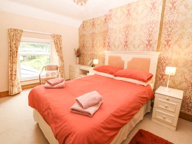 A bedroom with a double bed with red bedding bedside tables with lamps a window with floral curtains a chair and a desk with a vase and mirror at Dinas Farmhouse Annex in Caernarfon