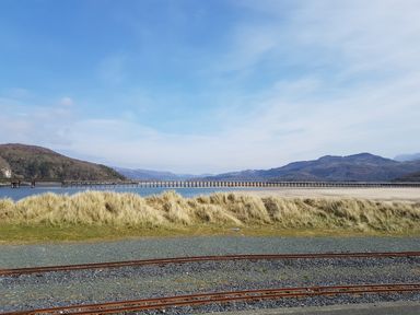 Railway tracks in front of grassy dunes with a long bridge over water and mountains in the background at Dehangfa in Fairbourne