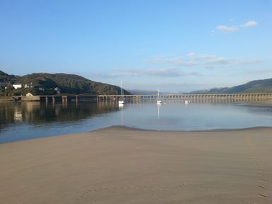 A sandy beach with footprints leading to calm water and boats near a bridge and hills at Dehangfa in Fairbourne