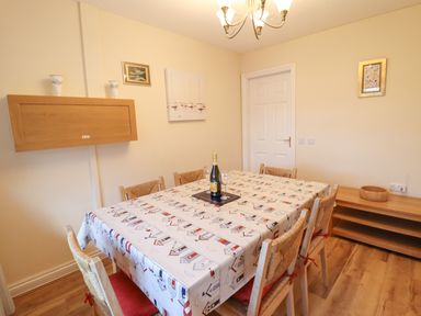 A dining room with a table covered by a patterned tablecloth and six chairs a bottle and two glasses on the table wooden shelves on the walls at Dehangfa in Fairbourne