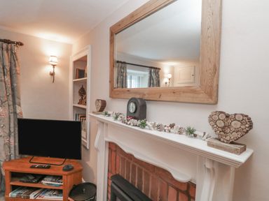 A room with a fireplace mantel decorated with pinecones and a heart-shaped wooden ornament a large mirror above the mantel a small television on a wooden stand with shelves a wall sconce light and floral curtains at Kirkstone Cottage in Ambleside