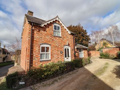 A brick house with white framed windows and a white door on a gravel driveway with bushes around at Elder Cottage in Mickleton near Chipping Campden