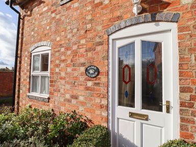Exterior view of a brick house with a white door featuring stained glass panels and a window with curtains at Elder Cottage in Mickleton near Chipping Campden