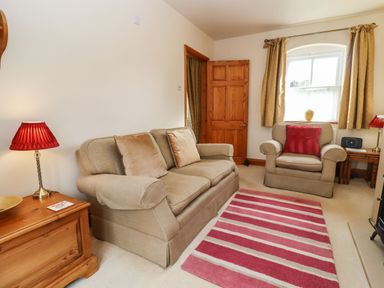 A living room with a beige sofa and armchair a striped red rug and a wooden cabinet with a red lamp at Elder Cottage Mickleton near Chipping Campden