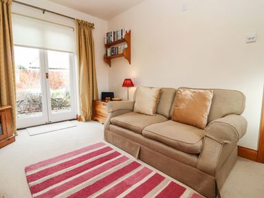 A sitting room with a beige sofa and two cushions a wooden side table with a lamp and bookshelves glass doors with curtains and a striped rug at Elder Cottage in Mickleton near Chipping Campden
