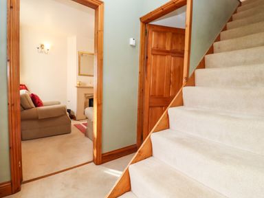 A hallway with carpeted stairs next to a doorway leading to a living room with armchairs and a fireplace at Elder Cottage in Mickleton near Chipping Campden