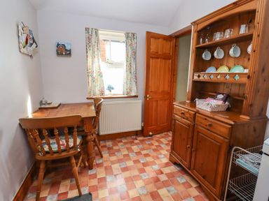 A small dining area with a wooden table and two chairs next to a window with floral curtains and a wooden cabinet with cups and plates at Elder Cottage Mickleton near Chipping Campden