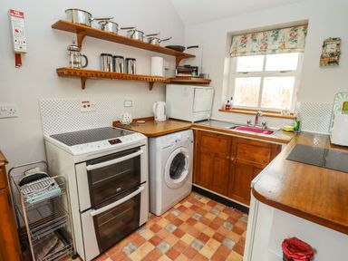 A kitchen with wooden shelves holding pots and containers a stove a washing machine a sink under a window and wooden cupboards at Elder Cottage in Mickleton near Chipping Campden