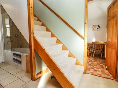 A staircase with carpet leading upstairs next to a bathroom with a bathtub and a towel and an open door showing a room with a wooden chair and table at Elder Cottage in Mickleton near Chipping Campden