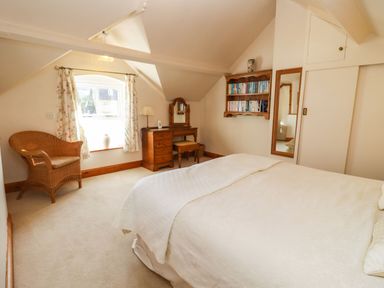 A bedroom with a bed a wooden dresser with mirror a wicker chair and a bookshelf at Elder Cottage in Mickleton near Chipping Campden