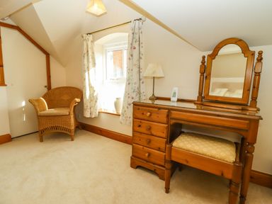 A room with a wicker chair next to a window and a wooden dressing table with a mirror and stool at Elder Cottage in Mickleton near Chipping Campden