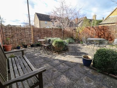 A patio garden area with wooden bench metal tables and chairs surrounded by plants and a wooden fence at Elder Cottage in Mickleton near Chipping Campden