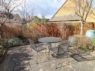 A patio with a round metal table and four metal chairs surrounded by bare trees and shrubs at Elder Cottage in Mickleton near Chipping Campden
