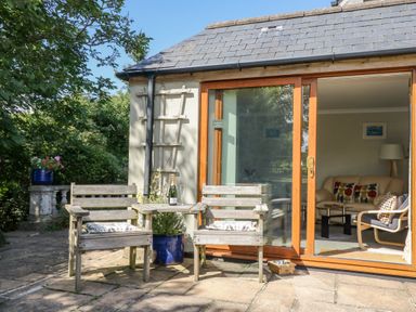 A patio with wooden chairs and a small table with a bottle and glasses outside a house with sliding glass doors showing a living room at Manege Cottage Near St Agnes