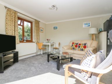 A living room with a beige sofa and patterned cushions a glass coffee table a TV on a stand and a small dining table with two chairs near the window at Manege Cottage Near St Agnes