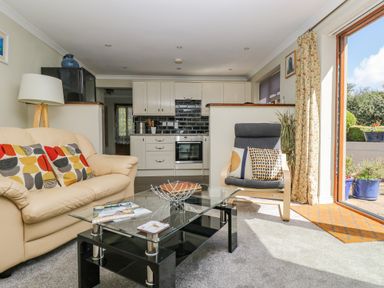 A living room with a beige sofa and patterned cushions a glass coffee table a grey armchair with two cushions and an open door to a garden at Manege Cottage Near St Agnes