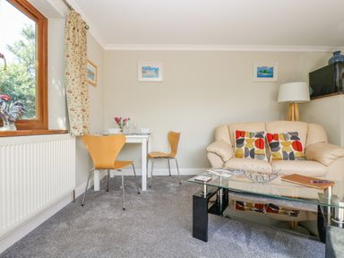A living room with a beige sofa with cushions a glass coffee table a small white dining table with two wooden chairs and a window with floral curtains at Manege Cottage Near St Agnes