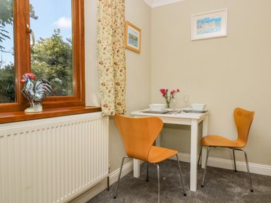 A small dining area with a white table set for two and wooden chairs near a window with a ceramic rooster on the sill at Manege Cottage Near St Agnes