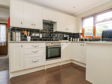 A kitchen with white cabinets black tile backsplash wooden floor and a window with a blind at Manege Cottage Near St Agnes