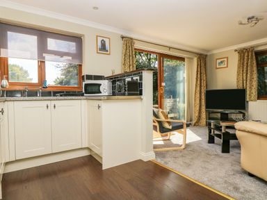 A kitchen with white cabinets and a microwave next to a living room with a TV and armchairs at Manege Cottage Near St Agnes