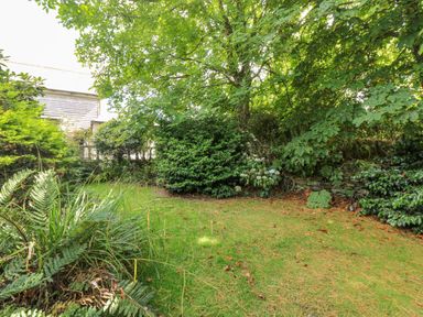 A garden with grass, bushes, a tree, and a stone wall near a wooden building at Manege Cottage Near St Agnes