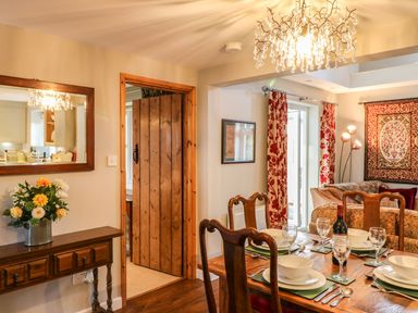 A dining area with a wooden table set for four with dishes and wine a wooden console table with flowers a wooden door and a living area with curtains and a wall tapestry at South Lodge in Hazelbury Bryan