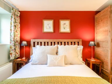 A bedroom with a wooden bed frame two lamps floral curtains and framed pictures on a red wall at South Lodge in Hazelbury Bryan