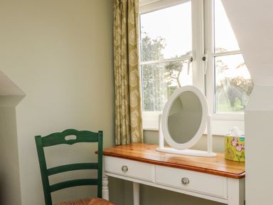 A room corner with a white dressing table holding a round mirror and a tissue box and a green wooden chair by the window at South Lodge in Hazelbury Bryan