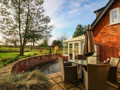 An outdoor patio with a round glass table and four wicker chairs next to a red brick building at South Lodge in Hazelbury Bryan
