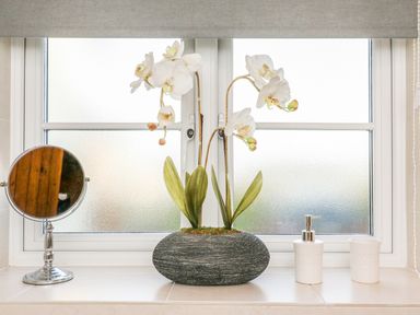 A bathroom window sill with a round mirror an orchid plant in a vase and two white containers at South Lodge in Hazelbury Bryan