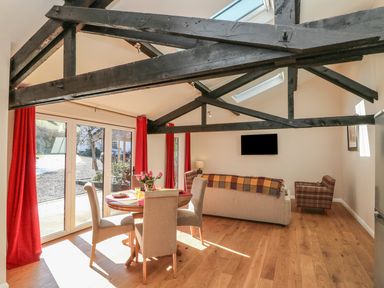 A living and dining area with a round table and four chairs next to glass doors with red curtains a sofa with a blanket a plaid armchair exposed ceiling beams and a wall-mounted television at Zeal Cottage in Crediton