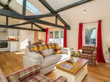 A living room with beige sofa, plaid armchairs, wooden coffee table, red curtains, and exposed black ceiling beams at Zeal Cottage in Crediton