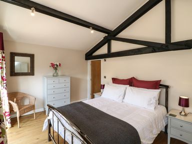 A bedroom with a metal bed frame white and red bedding a dresser with flowers a wicker chair and black ceiling beams at Zeal Cottage in Crediton