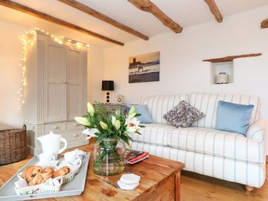 A living room with a striped sofa, wooden beams on the ceiling, a cabinet with string lights, a coffee table with a flower vase and a tea set at Polly's Bower in Landrake