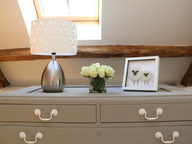 A gray dresser with white handles holding a table lamp with a patterned white shade a vase of white roses and a framed artwork of two sheep at Pollys Bower in Landrake