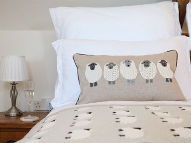 A bed with white pillows a beige cushion with five sheep illustrations a lamp and a glass on a wooden bedside table at Polly's Bower in Landrake