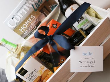 A gift basket with a black ribbon containing Cornish Fairings biscuits a bottle of Cornish pale ale a bottled drink a magazine and a note saying were so glad youre here at Polly's Bower in Landrake