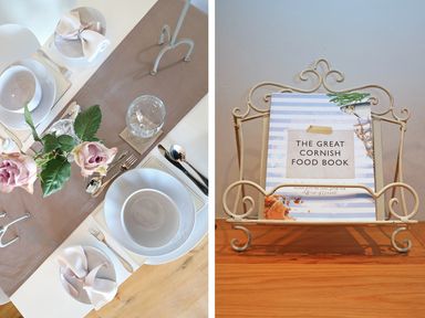 A dining table set with white plates, bowls, silverware, wine glasses, and pink roses alongside a book titled The Great Cornish Food Book in a decorative metal stand at Polly's Bower in Landrake