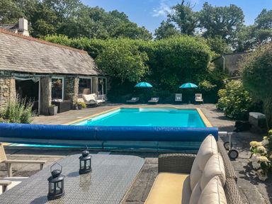 An outdoor swimming pool with lounge chairs and umbrellas beside a stone building at Polly's Bower in Landrake