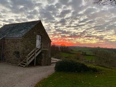 A stone building with stairs outside on a gravel path and green fields under a cloudy sunset sky at Polly's Bower in Landrake