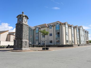 A stone clock tower on a roundabout with a tree and residential buildings behind at Y Enciliad in Rhosneigr