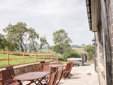 An outdoor patio with wooden tables and chairs next to a stone wall and a hot tub overlooking a grassy field with trees at The Farm in Presteigne