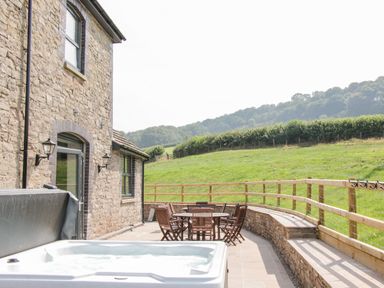 An outdoor patio with a hot tub wooden table and chairs by a stone house and fenced grassy hill at The Farm in Presteigne