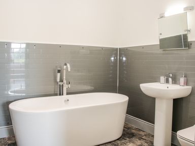 A bathroom with a white bathtub, sink, faucet, mirror, and grey tiled walls at The Farm in Presteigne