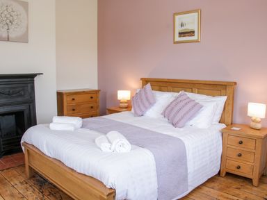 A bedroom with a wooden bed frame white bedding purple pillows and throws bedside tables with lamps and a black fireplace at The Farm in Presteigne