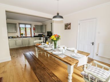 A dining area with a wooden table set with dishes and flowers next to a kitchen with cabinets and a window at Oakwood House in Rhayader