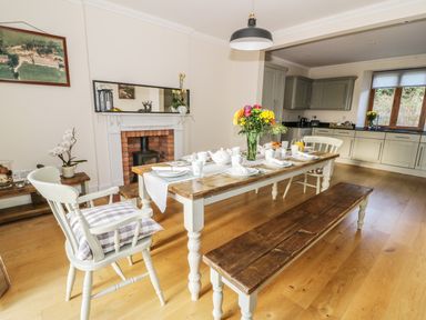 A dining area with a wooden table set for breakfast and chairs next to a fireplace and kitchen at Oakwood House in Rhayader