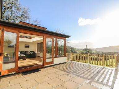 An outdoor patio with wooden railing overlooking hills and an interior living room with sofas and a coffee table at Oakwood House in Rhayader
