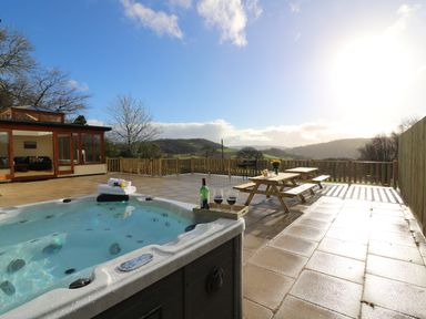 An outdoor patio with a hot tub, wooden picnic tables, and a view of hills at Oakwood House in Rhayader