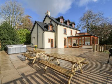 An outdoor patio with wooden picnic tables a hot tub and a large house with multiple windows and a glass extension at Oakwood House in Rhayader